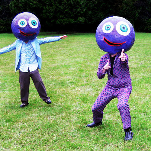 A photo of Otis Fodder and Mildred Pit standing in a field, wearing big purple smiling masks. One is wearing a light blue suit and the other a purple one with a diamond pattern.